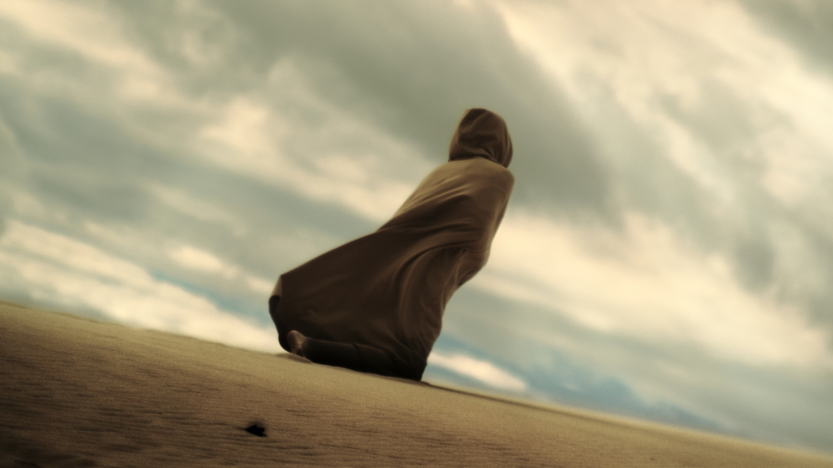 Person in a brown coat sitting on a sand dune with a dramatic sky.